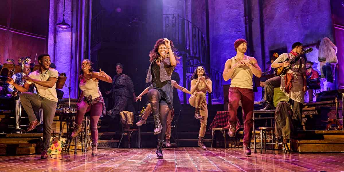 Bethany Antonia and the cast of Hadestown perform energetically on stage, surrounded by vibrant lighting and set pieces.