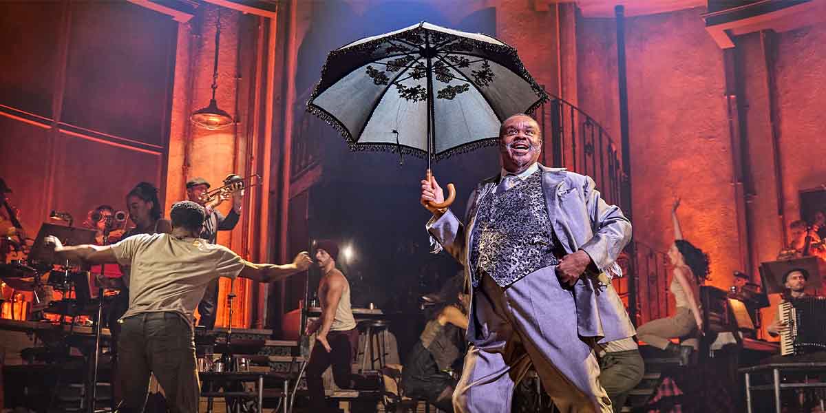 Clive Rowe smiles while holding a decorative umbrella, surrounded by performers in a vibrant theatre setting.