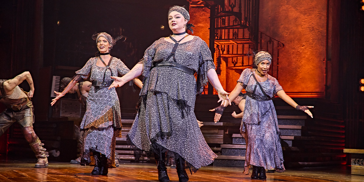 Four performers in period costumes, singing on stage with dramatic lighting and a dark, atmospheric backdrop.