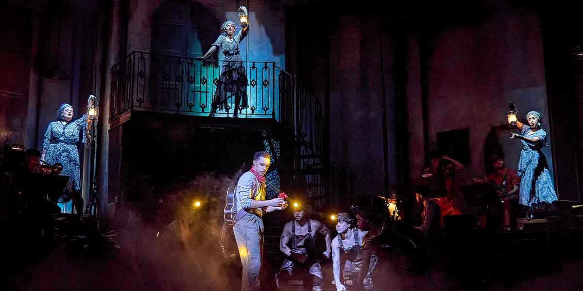 Spike Maxwell holds a lantern while surrounded by cast members in a dramatic, smoky stage setting.