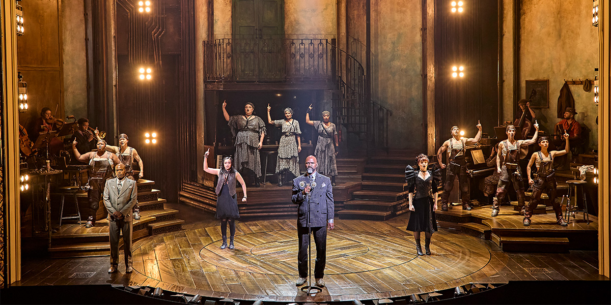 The cast of Hadestown poses together, wearing period costumes with rich textures, against a dimly lit stage backdrop.