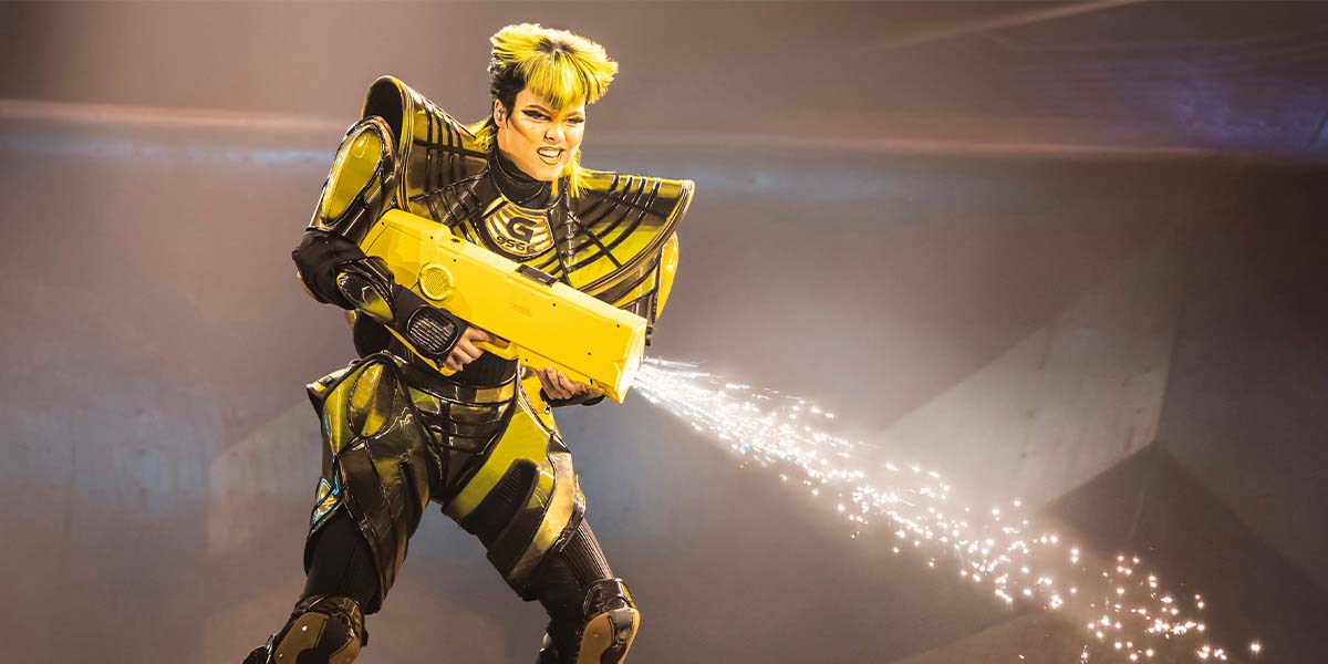 A performer in a yellow and black costume sprays sparks from a yellow device, smiling energetically.