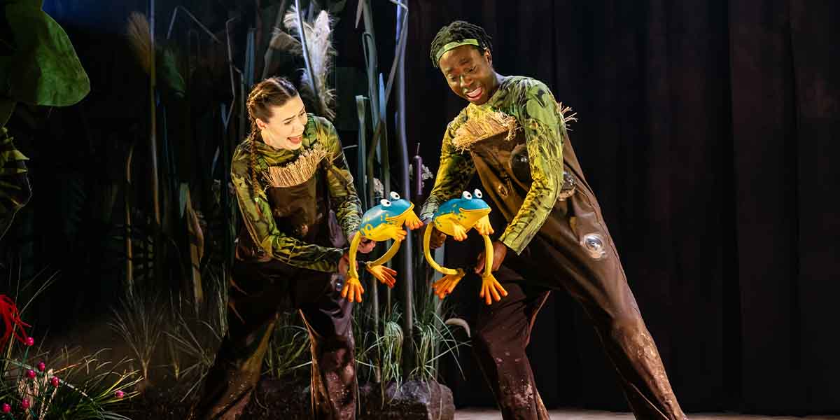 Ciara Hudson and Jordan Eskeisa perform with frog props, wearing green costumes in a vibrant stage setting.