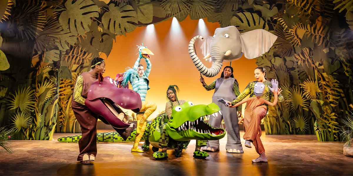 Actors in vibrant animal costumes perform on stage, surrounded by a jungle-themed backdrop with bright lighting.