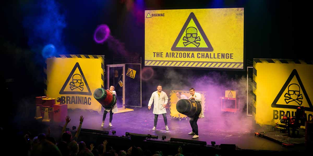 Three performers in lab coats hold large cannons on stage, with vibrant smoke and a large screen backdrop.