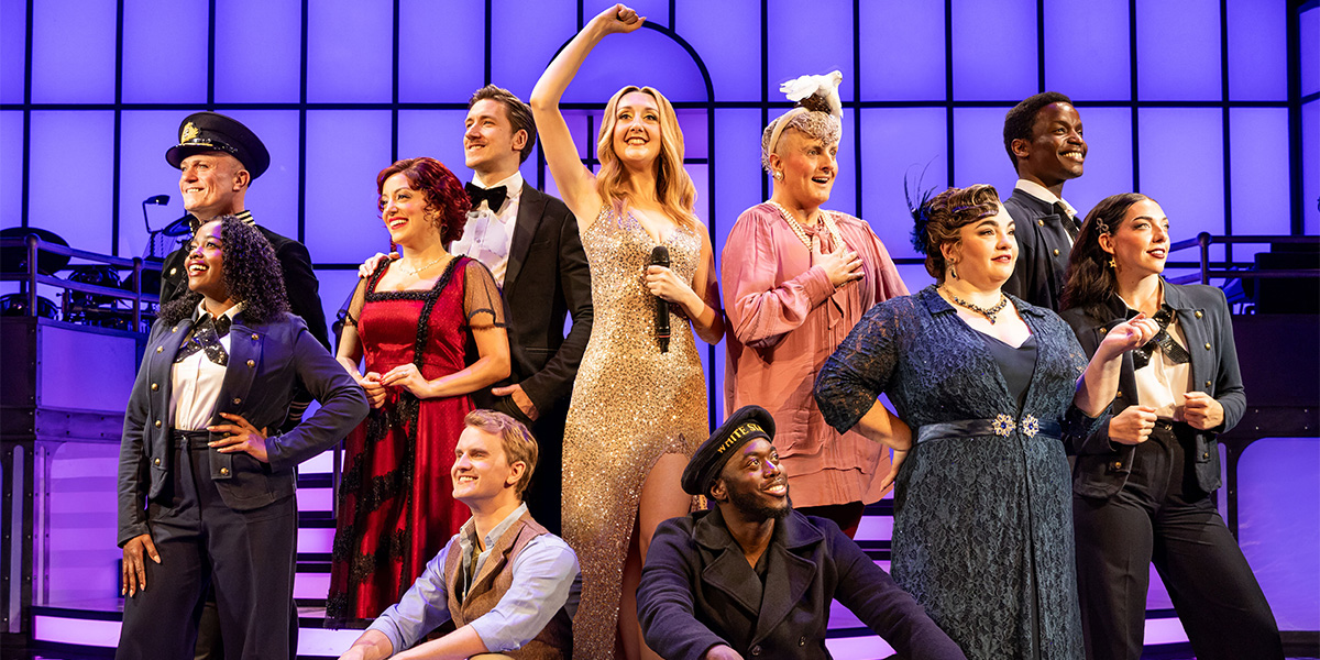 Cast members of varying ages and outfits, standing on a stage with a vibrant purple backdrop, enthusiastically posing during a performance.