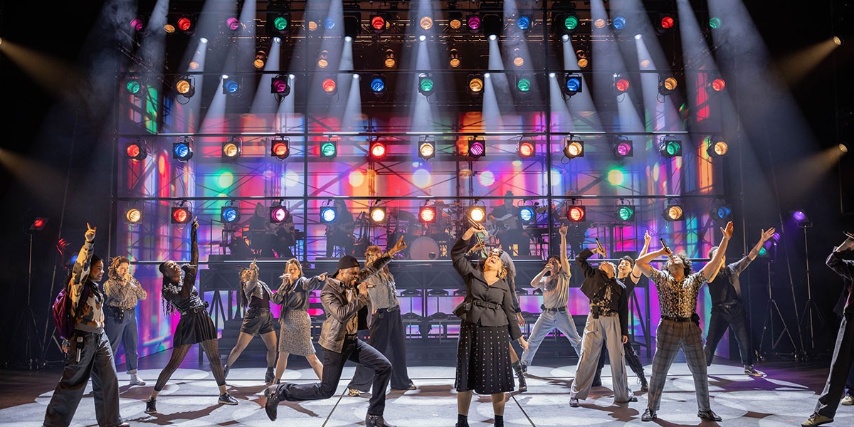 A performer in a vibrant costume sings passionately on stage, with bright lights and an enthusiastic audience behind.