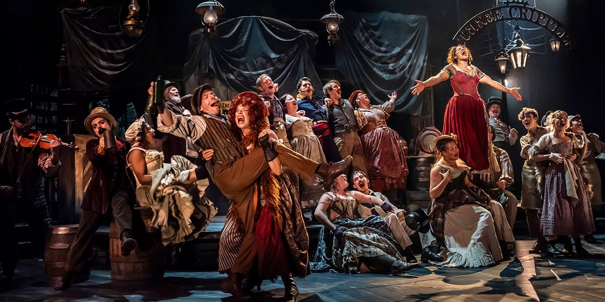 A performer in a vibrant costume poses dramatically on stage, with colourful lighting and a theatrical backdrop.