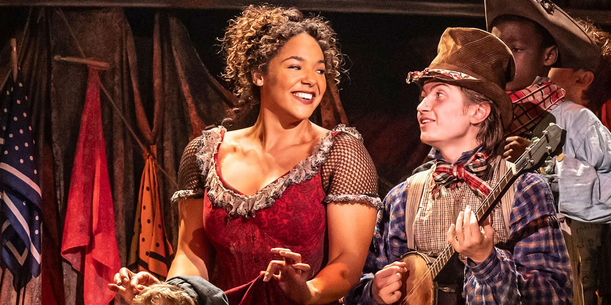 Three actors in period costumes perform on stage, with vibrant lighting and a theatrical backdrop.