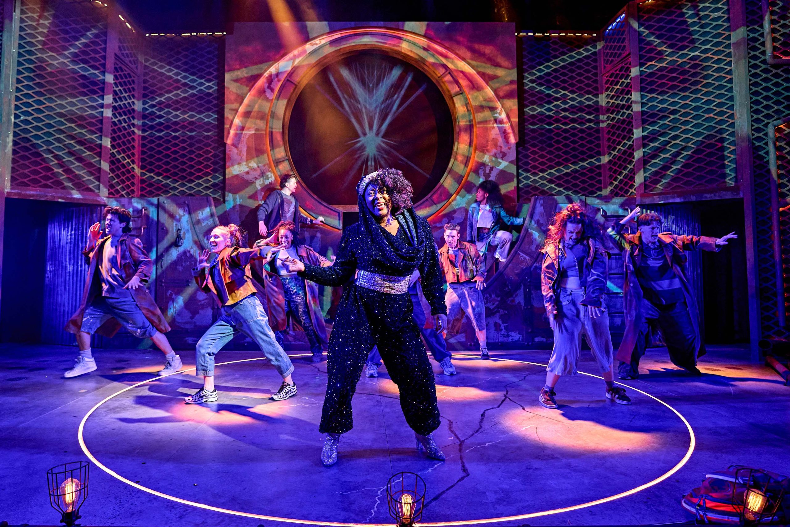 The central figure is dressed in a dramatic, textured, black shaggy costume with a large hood and sunglasses, exuding a powerful and mysterious presence. They are confidently posing or gesturing, drawing attention to themselves. In the background, other performers are visible, some partially covered in translucent fabric that resembles veils or ethereal cloaks, contributing to the surreal and mystical aesthetic. The set design includes curved structures with abstract, colorful patterns. The overall atmosphere is magical and otherworldly, suggesting a fantasy or mythological theme.