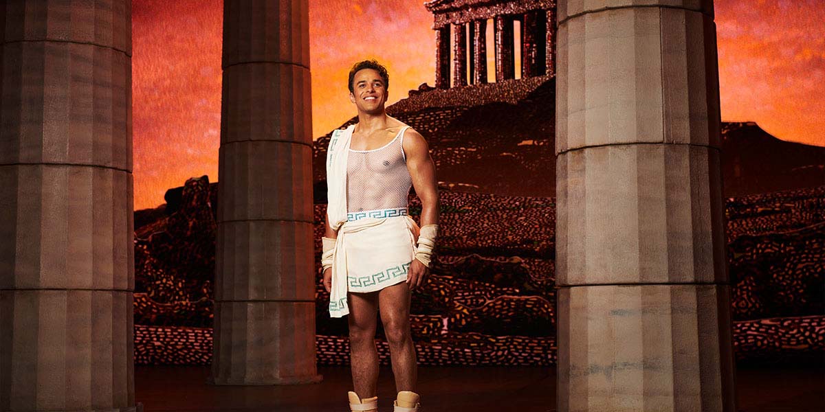 Luke Brady as Hercules poses heroically, wearing a blue tunic and brown sandals, against a vibrant backdrop.