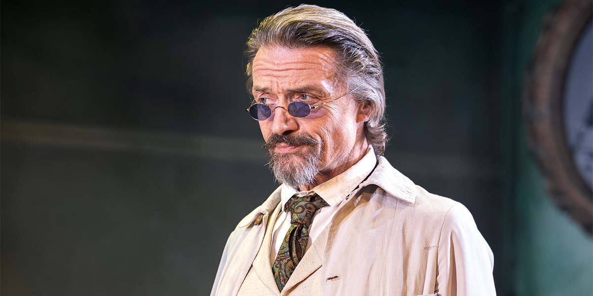 A man with grey hair and glasses, wearing a beige jacket, stands in a dimly lit setting.