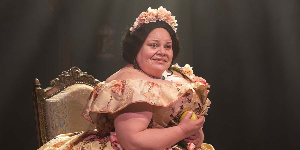A woman in a floral gown sits on a chair, holding a lemon, with soft lighting in the background.