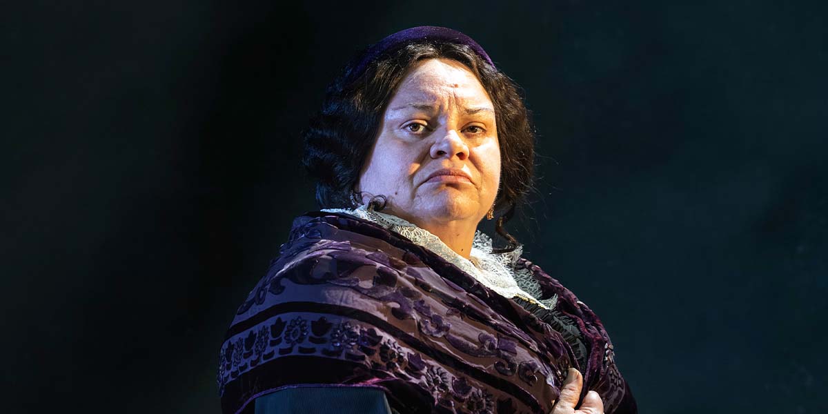 A woman in a dark purple shawl looks thoughtfully at the camera, with a textured black background.