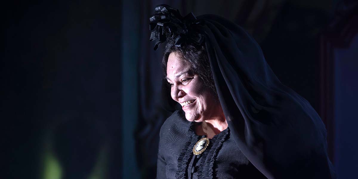 A woman in a black dress and veil smiles, illuminated by soft lighting against a dark background.
