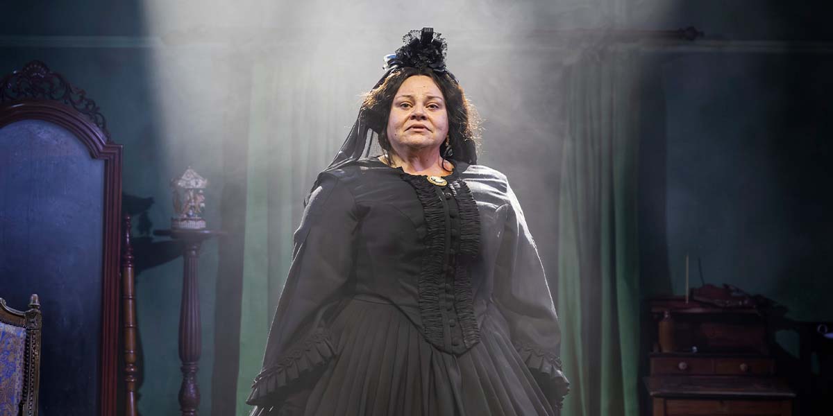 A woman in a black Victorian dress stands confidently, surrounded by soft lighting and green curtains.