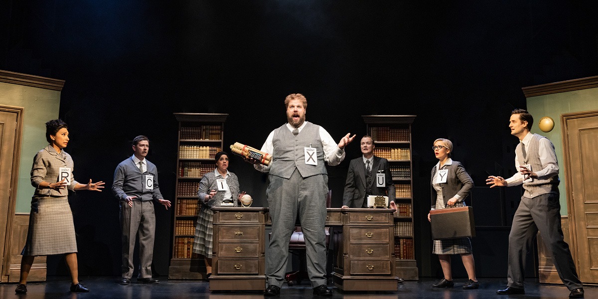 The company of The Comedy About Spies are standing around Agent X who is holding n explosive.