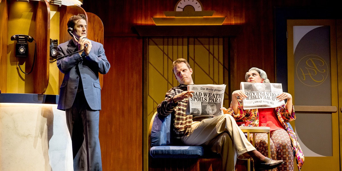 two people sit on chairs, while another behind them is on the telephone. Those sitting, appear to be listening in to the telephone conversation.