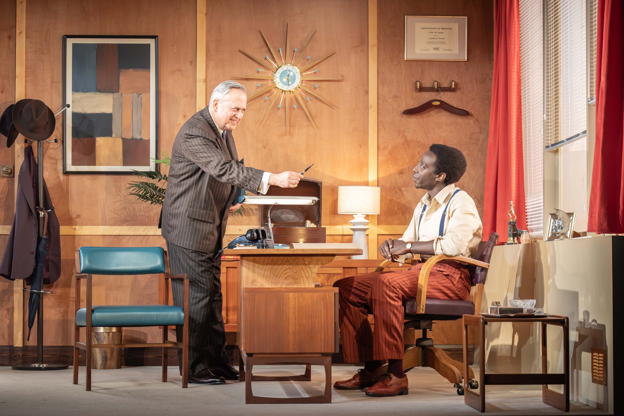 On the left a person wearing a white shirt and brown trousers sits at a desk. On the right a person wearing a brown suit stands facing them holding a pen out towards them.