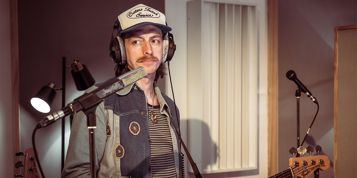 A musician plays a guitar on stage, illuminated by colourful lights, with a cheering audience in the background.