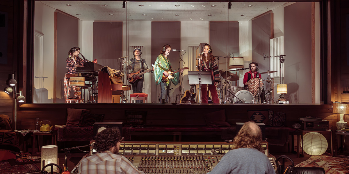 A musician plays a guitar on stage, illuminated by colourful lights, with a cheering audience in the background.