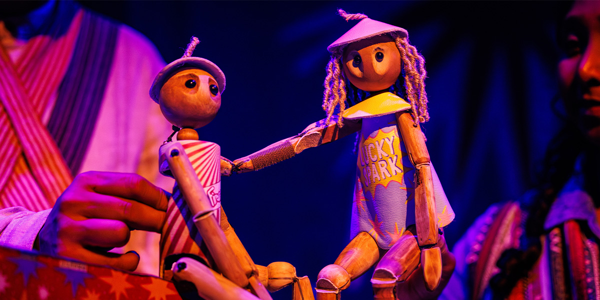 Two wooden puppets are on stage under warm purple lighting.