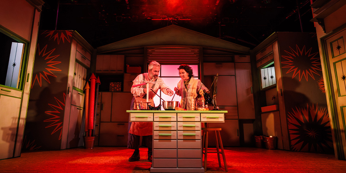 A young girl in a vibrant costume stands on stage, surrounded by colourful fireworks and dramatic lighting.