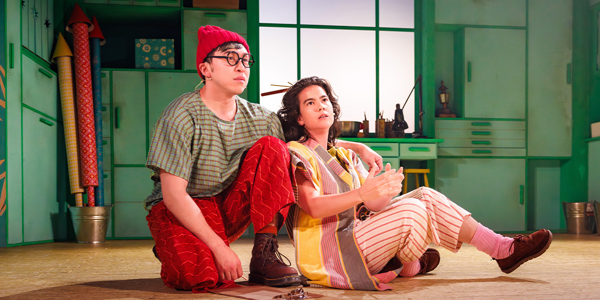 Two people in vibrant costume sit side by side on stage.