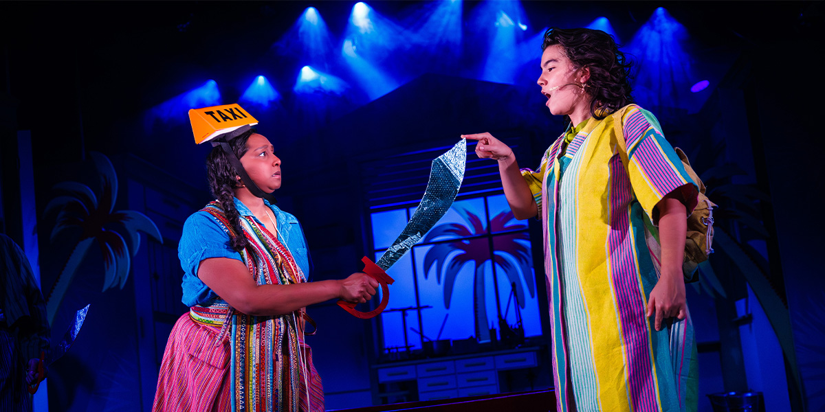 A scene from "The Firework Maker's Daughter" featuring two actors in vibrant costumes. One wearing a taxi sign on their head.