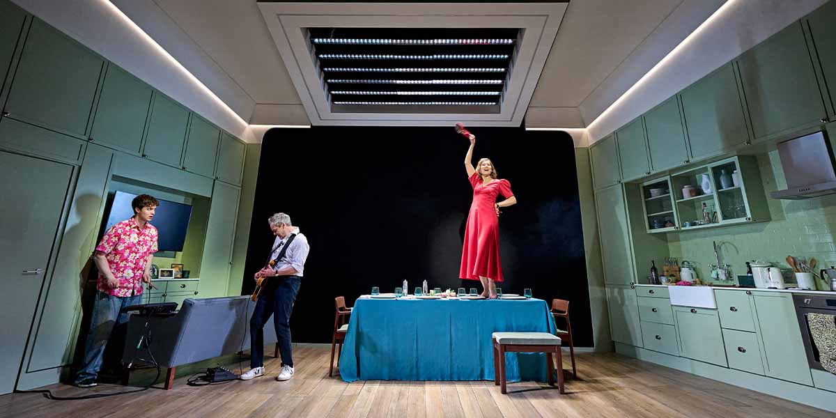 Rosamund Pike, in a red dress, stands on a table, while Jamie Glover plays guitar nearby.