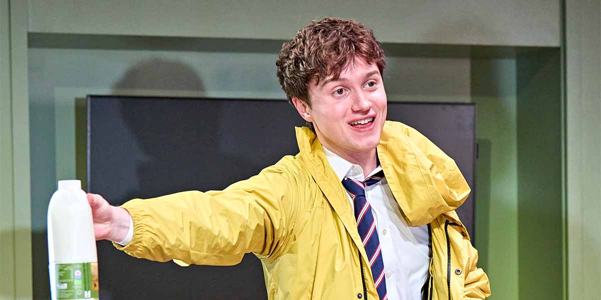 Cormac McAlinden, wearing a yellow jacket, smiles while holding a bottle in a theatrical setting.
