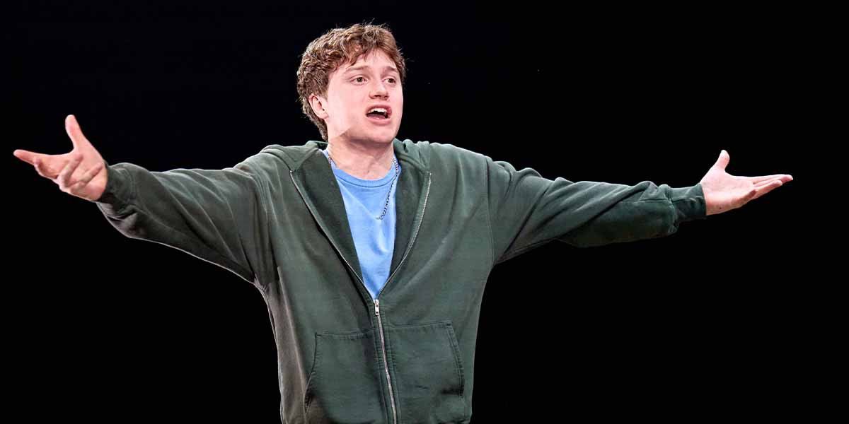 Cormac McAlinden, wearing a green hoodie, gestures expressively while performing on stage against a dark background.