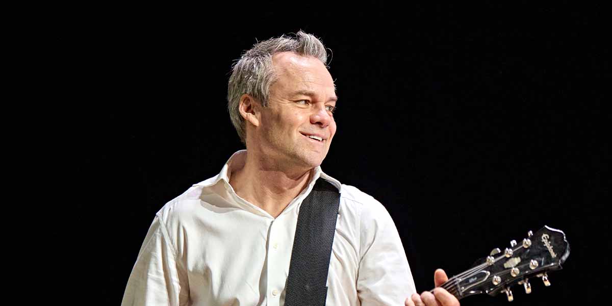 Jamie Glover smiles while holding a guitar, wearing a white shirt against a dark background.