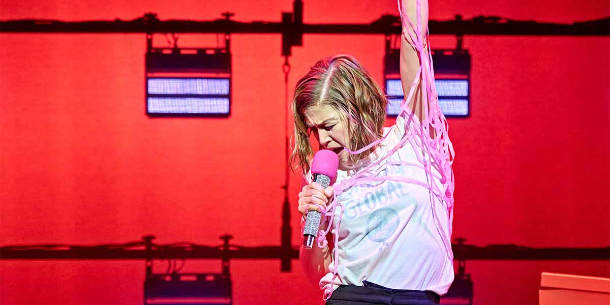 Rosamund Pike, as Jessica, performs passionately on stage, holding a microphone with vibrant red lighting behind her.