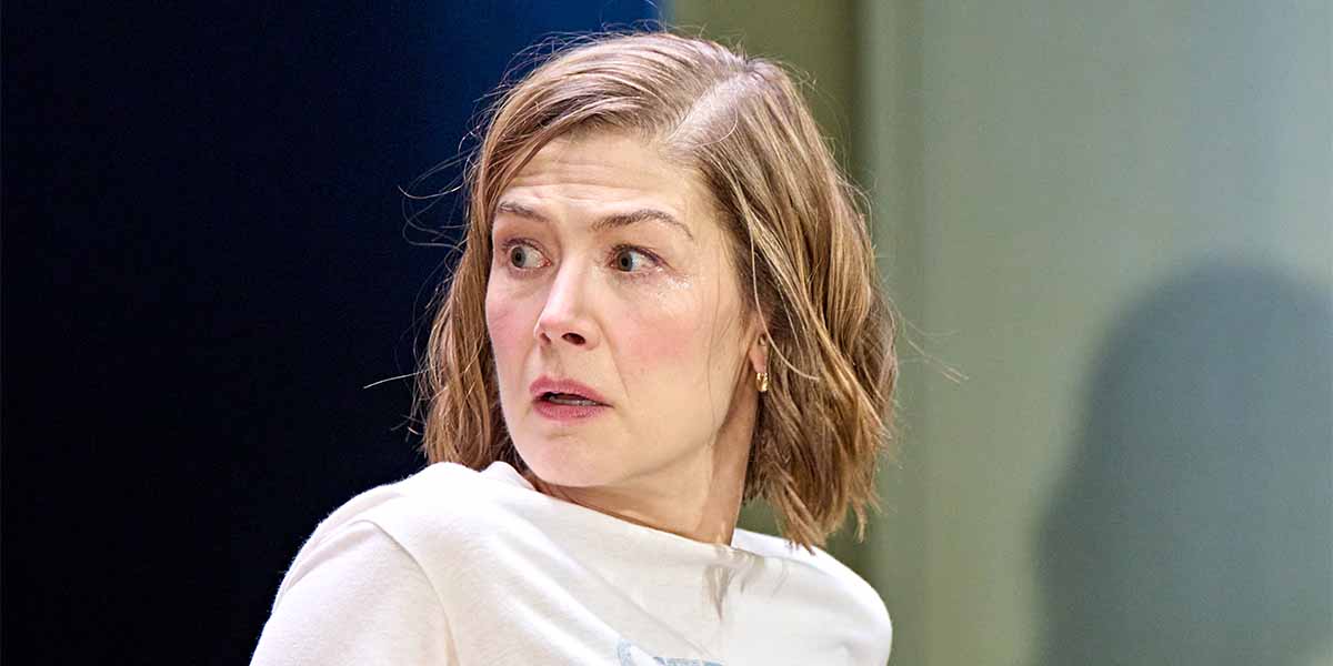 Rosamund Pike, portraying Jessica, looks startled with short hair, wearing a light-coloured top, against a dark background.