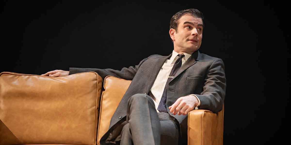 Arthur Wilson sits on a brown leather sofa, wearing a grey suit and tie, looking thoughtfully to the side.