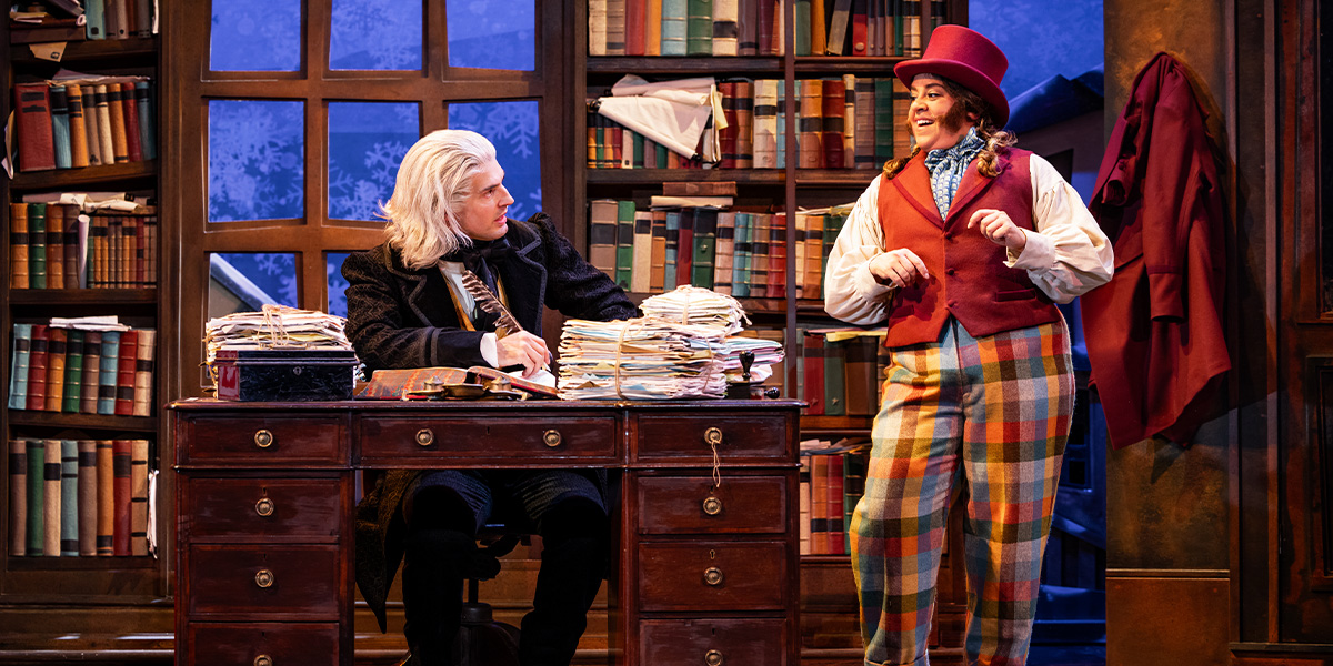 Daniel Fraser and Nancy Zamit pose together, smiling, in festive costumes with Christmas decorations in the background.