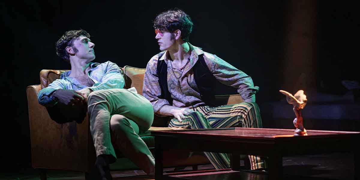 Calam Lynch and Noah Ritter sit on a sofa, engaged in conversation, with a wooden table nearby.