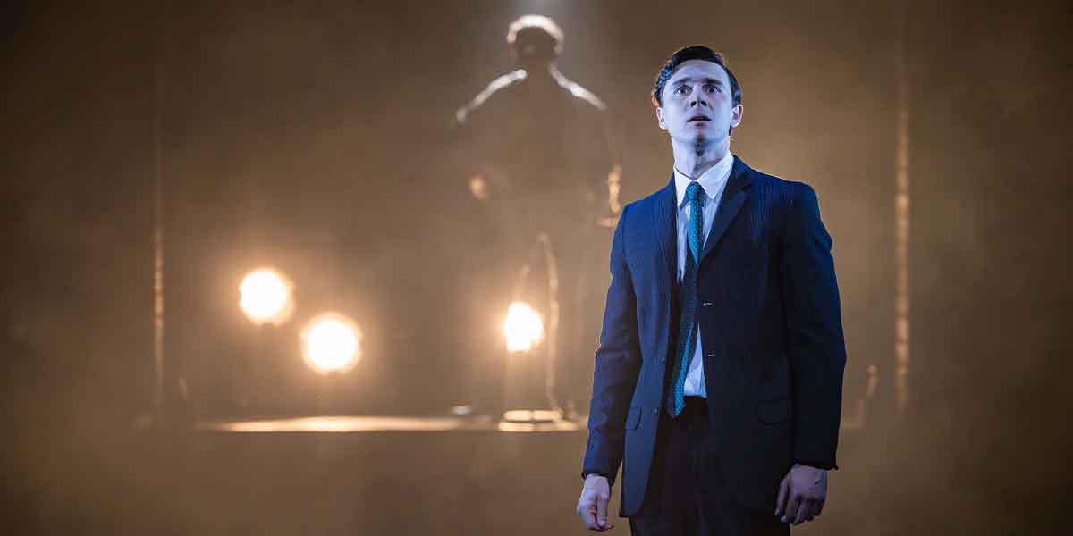 Calam Lynch, dressed in a suit, gazes upwards against a smoky background with bright stage lights.