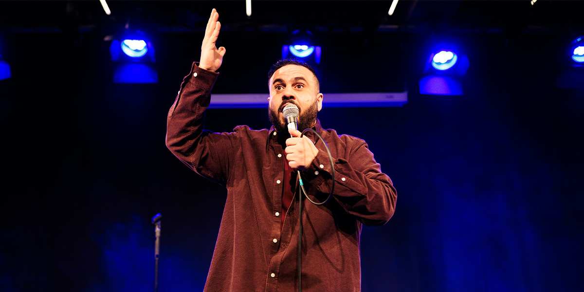 A performer with a beard performs on stage, gesturing with one hand, under blue stage lights.