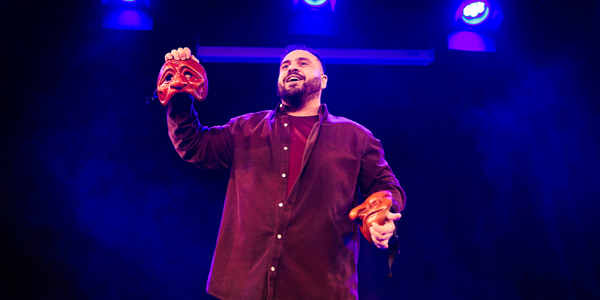 A performer holds two red masks, smiling under blue stage lights with a dark background.