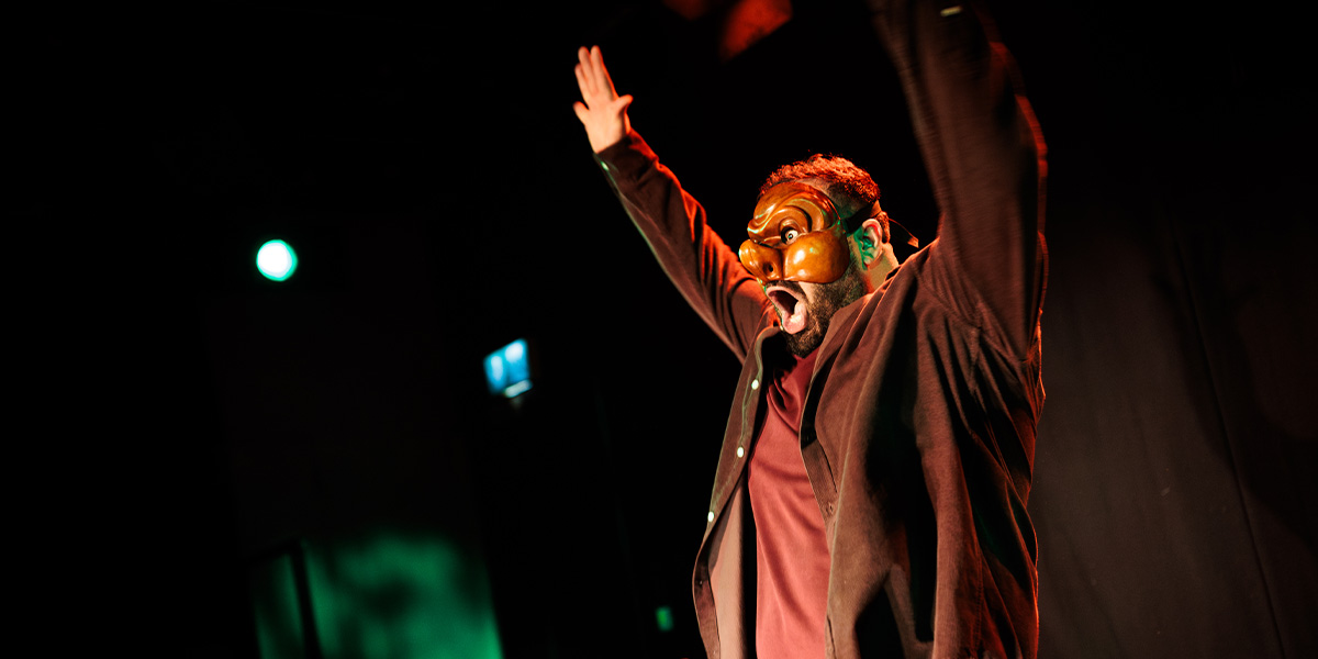 A performer wearing a brown mask raises his arms dramatically against a dark background with green lighting.