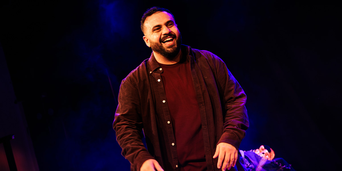 A smiling performer in a dark shirt and jacket performs on stage, with blue lighting in the background.