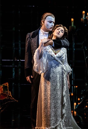 A masked figure in a black cape stands in a dimly lit theatre, with ornate decor in the background.