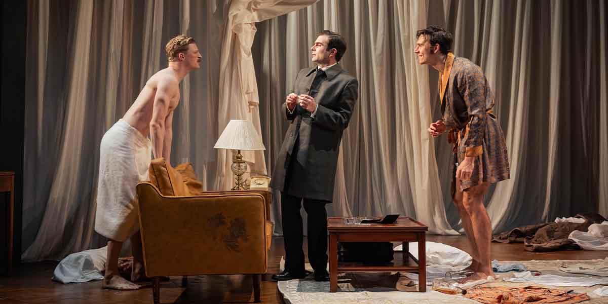 Three people interact in a messy room; one is partially clothed, while another wears a robe.