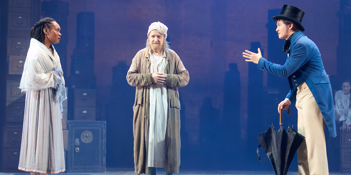 A scene from A Christmas Carol featuring actors in Victorian costumes, with festive decorations in the background.