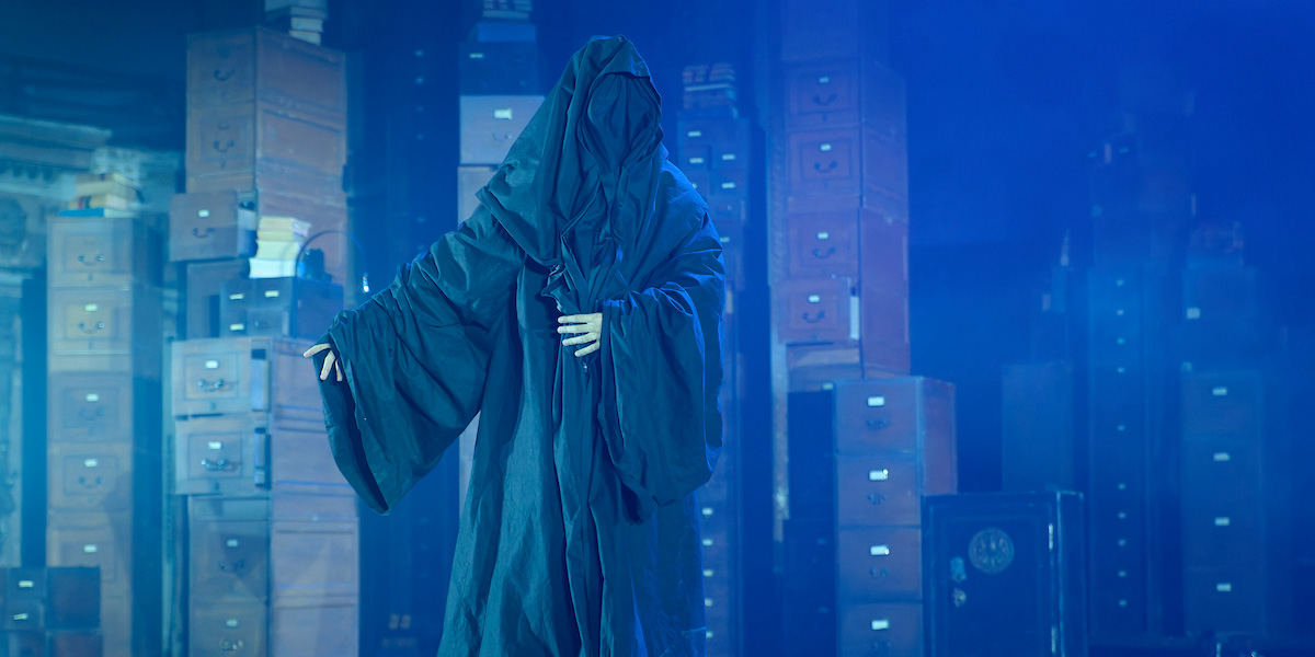 A cloaked figure stands with outstretched arms against a backdrop of filing cabinets and blue lighting.