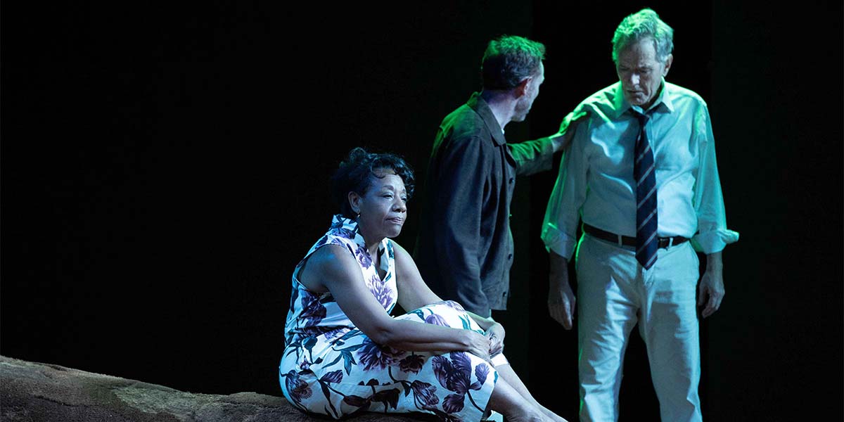 Marianne Jean-Baptiste as Kate Keller sits on a rock, while Richard Hansell and Bryan Cranston converse nearby.