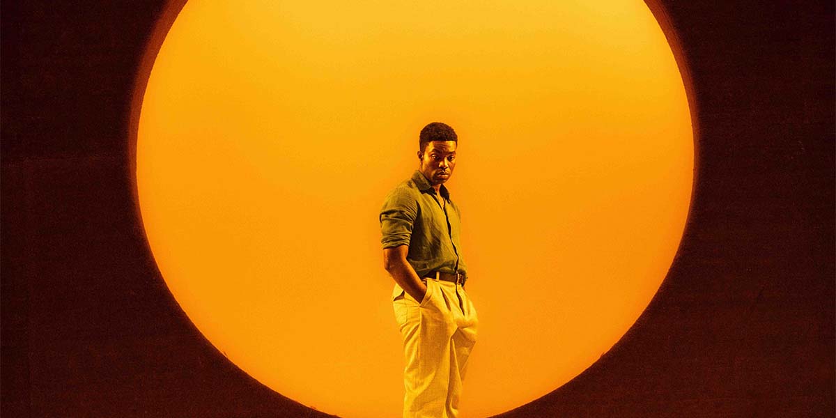Paapa Essiedu stands confidently in front of a large, glowing orange circle, wearing a green shirt and beige trousers.