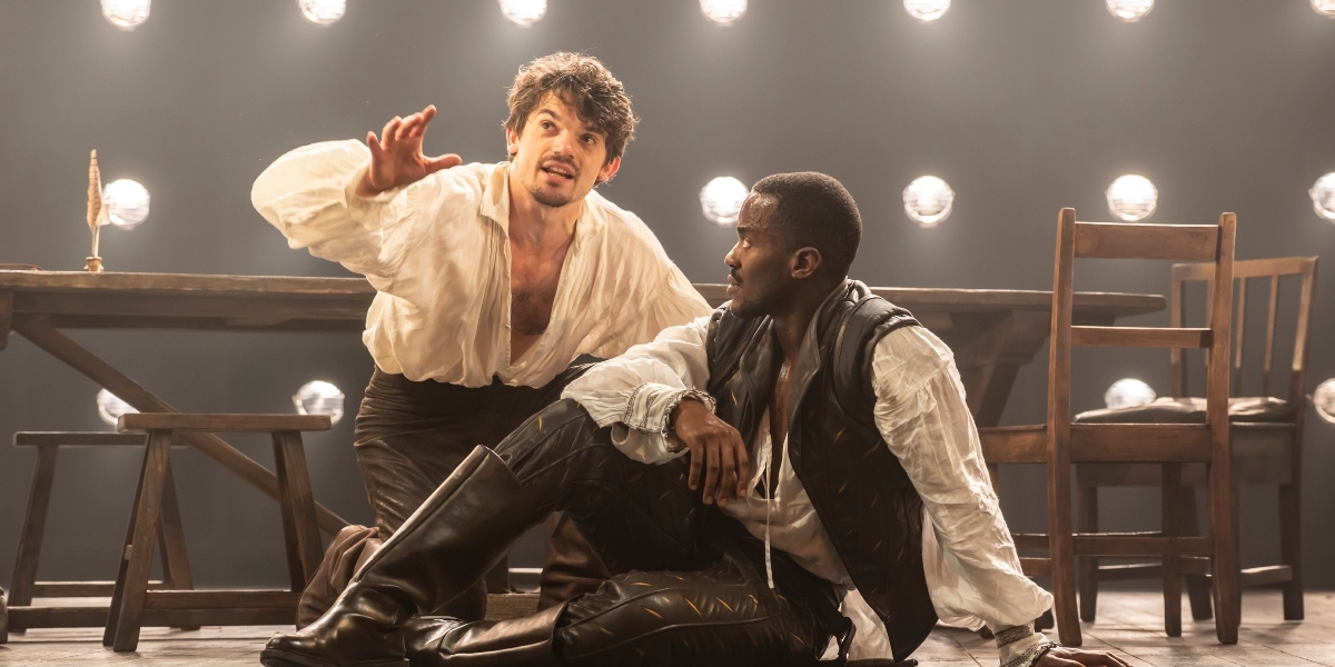 Two actors in period costumes perform on stage; one kneels with an arm raised and an intense expression, while the other sits on the floor looking up at him. Wooden chairs, a table, and round stage lights are in the background.